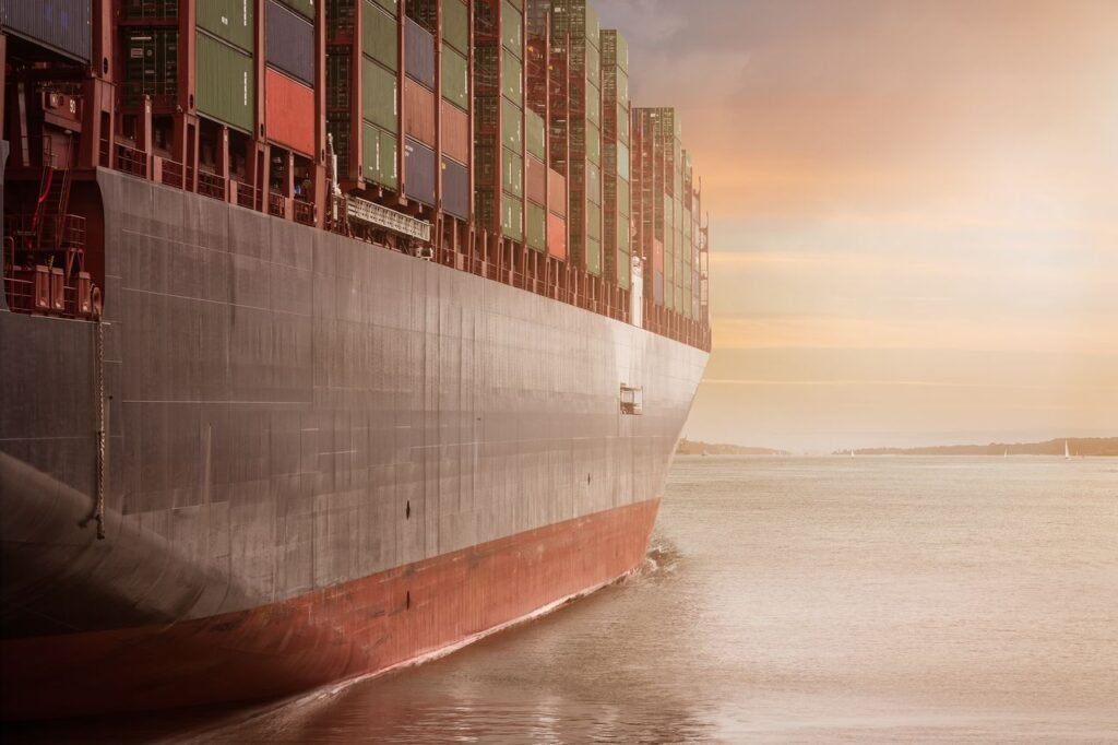 الرئيسية A colossal cargo ship loaded with containers navigates through calm waters against a vibrant sunset sky.